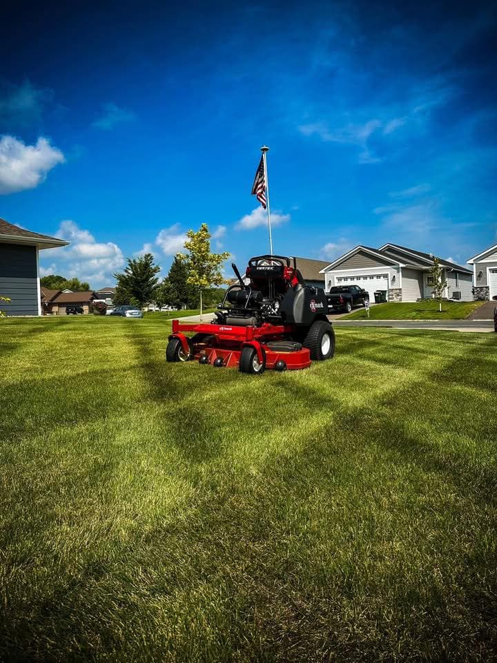 Lawn mower with American flag cutting grass in residential neighborhood under blue sky.