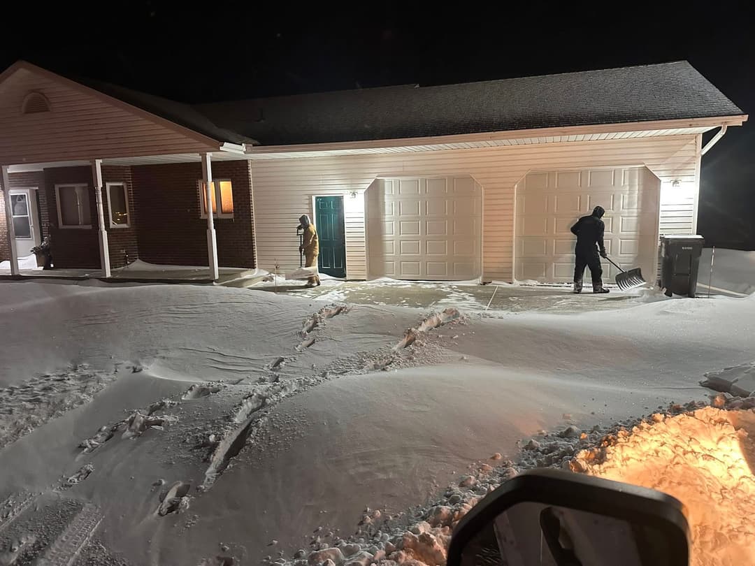 People shoveling snow near a house and garages at night during a winter storm.