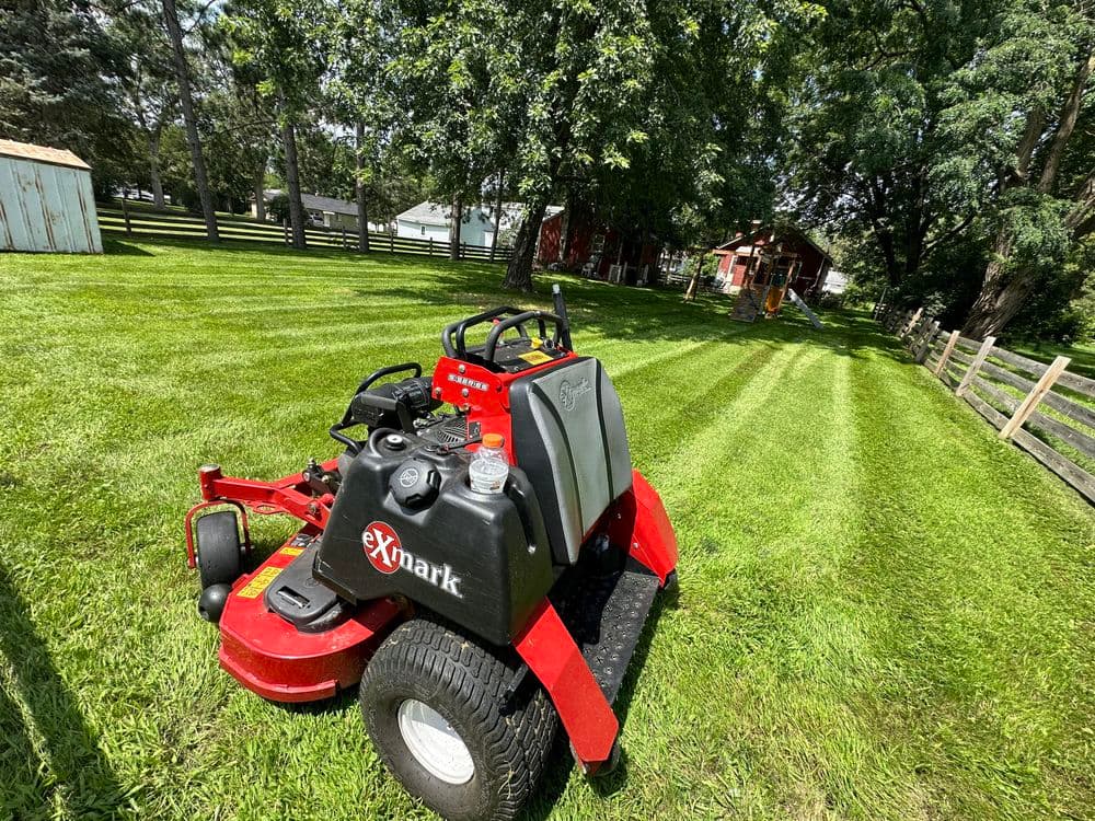 Exmark lawn mower on freshly mowed grass with trees and buildings in the background.
